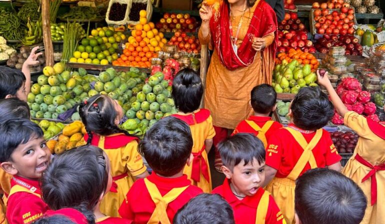 A group of preschool children in red and yellow uniforms visit a fruit market, listening to a teacher explaining about fruits.