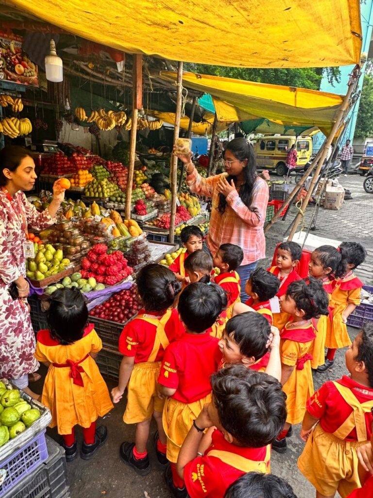 A group of preschool activates in red and yellow uniforms visit a fruit market, listening to a teacher explaining about fruits.